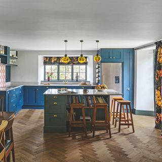country kitchen ideas, country kitchen with bright blue units, sage green island, yellow glass pendant lights, herringbone floor, patterned floral curtains and blind, bar stools, white worktops
