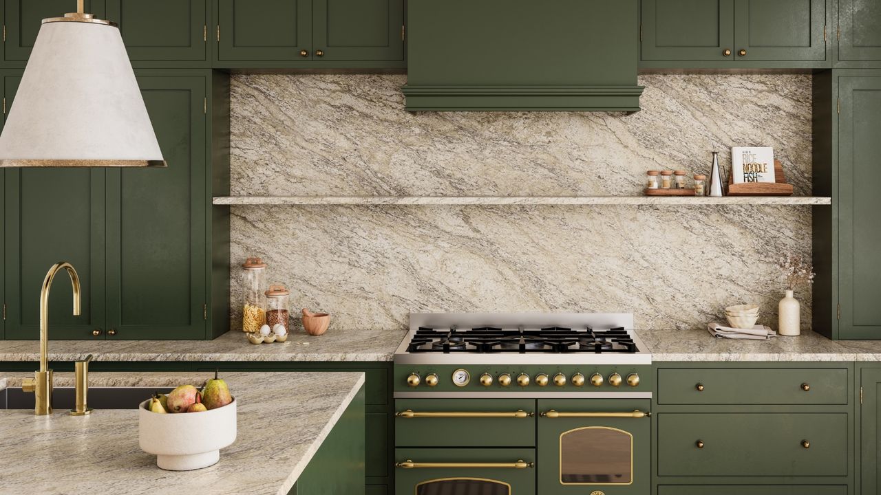 Modern, cohesive green kitchen with marble countertops, and floating shelves surrounded by green cabinets. A bowl of fruit is pictured on a kitchen island in the foreground, below a white lampshade.
