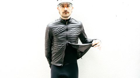 A white man wears a thin balck insulated cycling jacket against a white wall