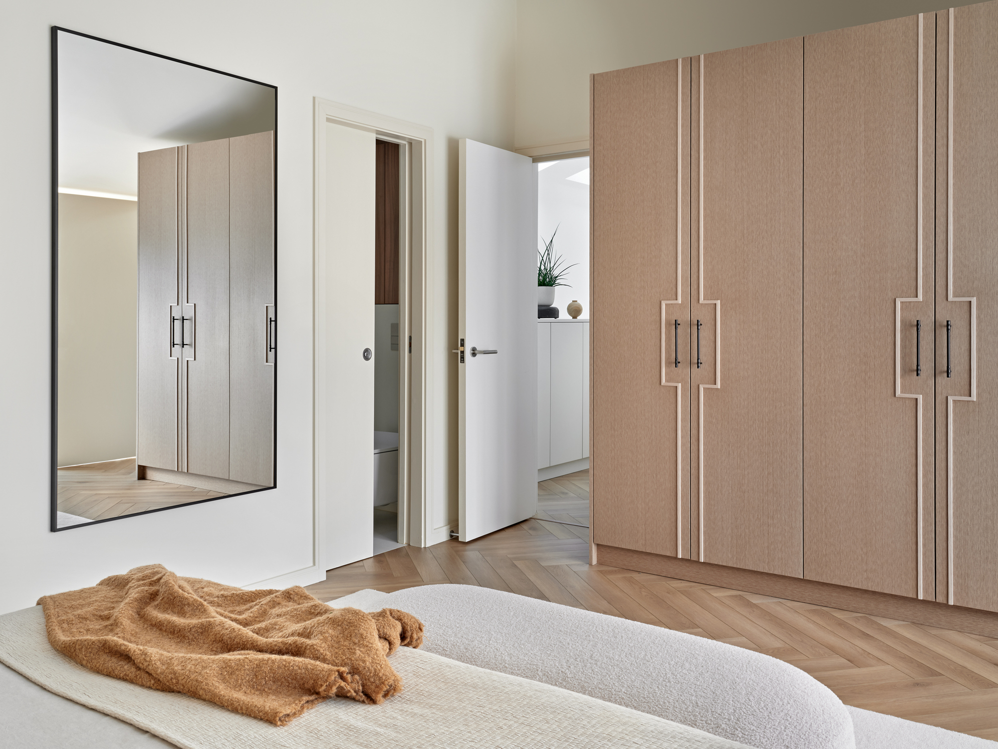 modern bedroom with herringbone floors, wall-mounted mirror, white bedding with yellow throw draped across the end of the bed, upholstered bench seat at the foot of teh bed, and built-in timber wardrobes