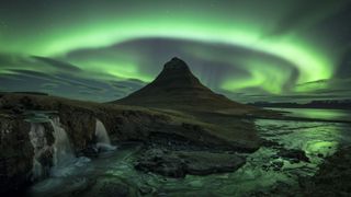 northern lights appear as a large green band swirling over the top of the central mountainous peak reflected in the water below.