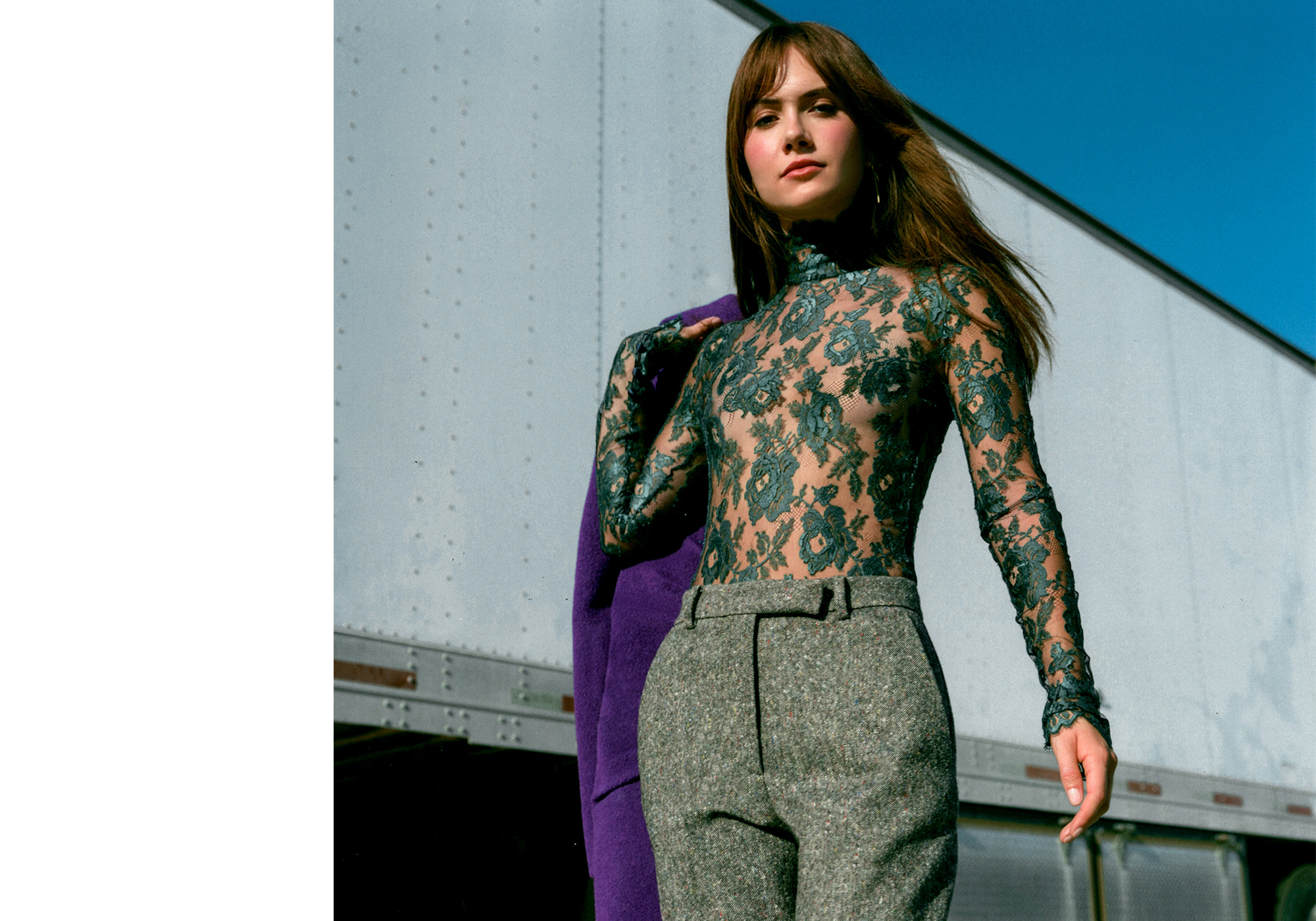 Emilia Jones for WWW's fall Ones to Watch portfolio. She is standing next to a white semi truck holding a purple coat and wearing a green lace high-neck top and gray trousers.