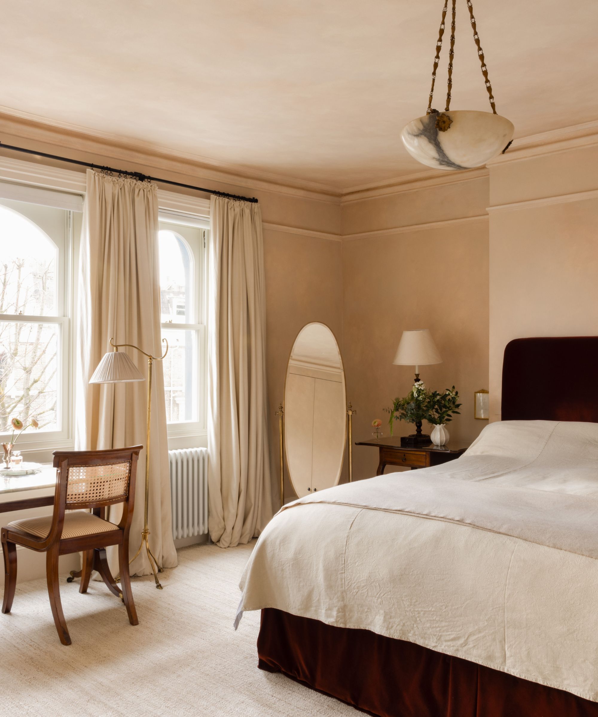 Bedroom with plaster walls, dark red velvet bed with linen bedding, round floor mirror, a vintage desk with a wooden chair and floor lamp and an antique marble ceiling light