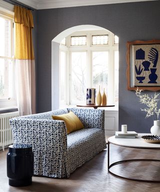 A small blue-gray living room with a compact square blue and white patterned sofa away from the walls, large windows with white and yellow drapes, a round coffee table and white, yellow and blue accessories.