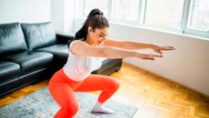 Woman squats in a living room, her knees are bent and her hips are pushed back, she holds her arms straight our in front of her. She is wearing bright orange athletic leggings, a white top and her black hair is in a high ponytail.