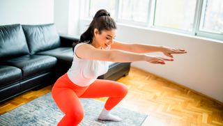 A woman in workout clothes performs a squat in a living room. She is bending at the knees and hips to move into the squat and holds her arms out in front of her. Behind her we see a sofa and window.