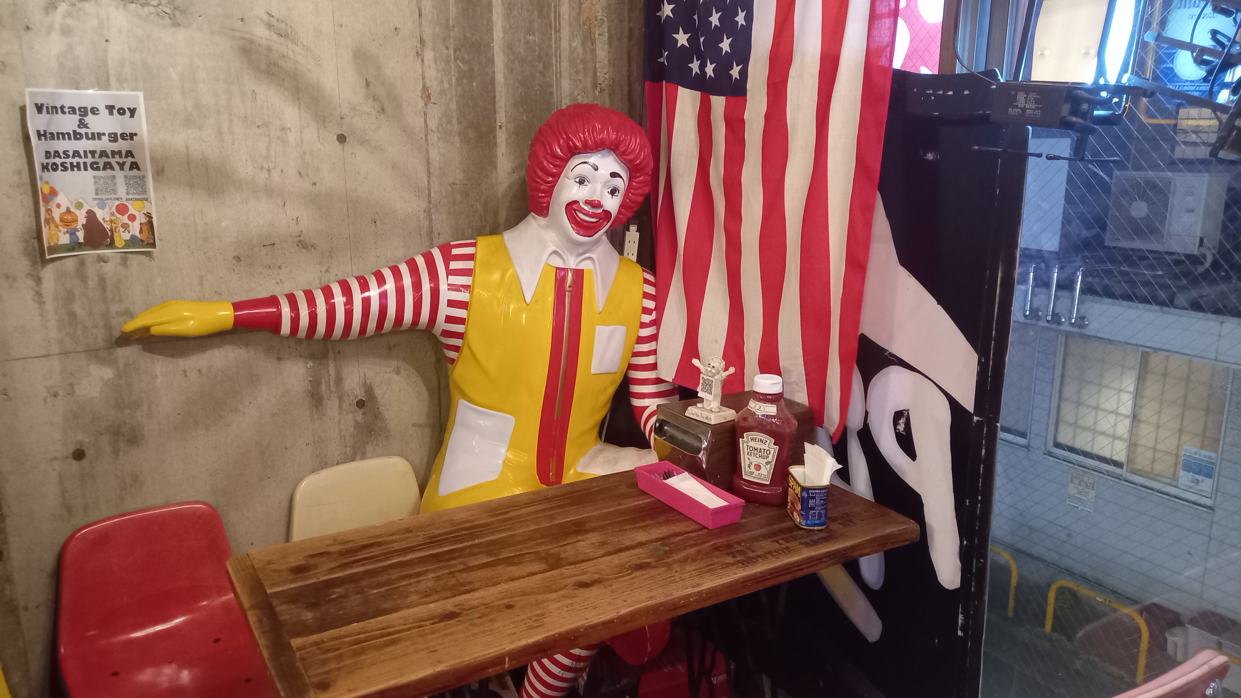 A life-size Ronald McDonald statue sits at a wooden diner table draped with an American flag, arms outstretched in welcome, inside the Vintage Toy &amp;amp; Hamburger restaurant in Koshigaya, Saitama.