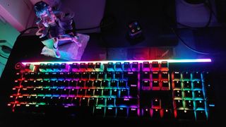 In a darkened room, Be Quiet's Light Mount gaming mechanical keyboard can be seen with its RGB lights on. The RGB lights are one of the only light sources in the dark room, illuminating a nearby Love Sailor Miku Hatsune anime figurine, and a fidget cube.