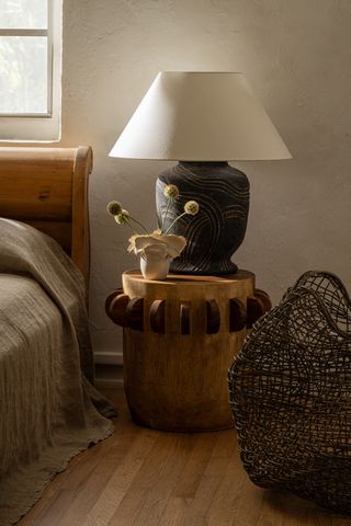 A lamp with a wide, white linen shade and roughly decorated black finish shits atop a wooden bedside table in a rustically decorated bedroom.