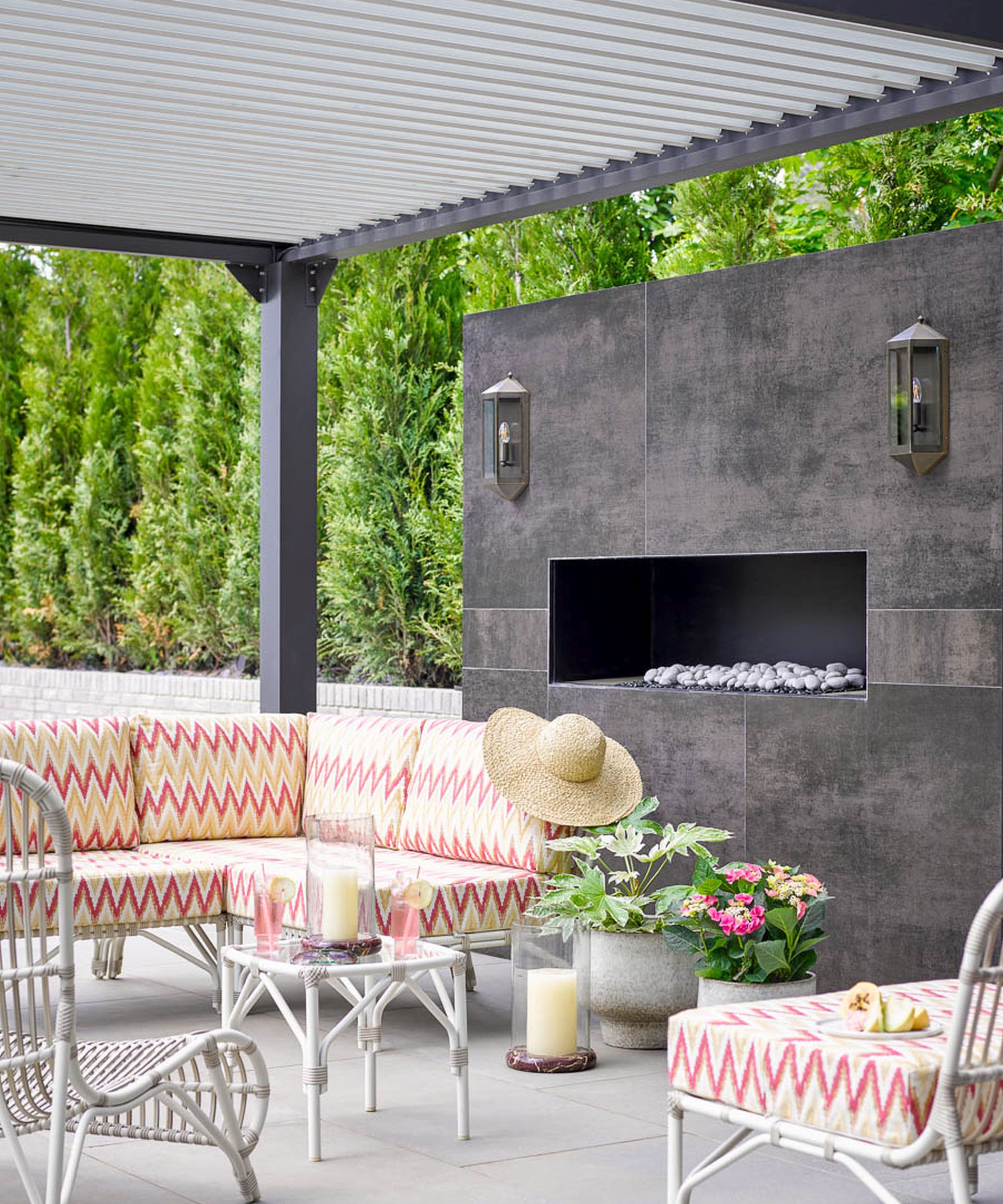 A modern outdoor lounge area under a gray pergola. It features a sectional sofa with red and yellow chevron cushions, a sleek dark gray fireplace wall with built-in lanterns, and white wicker accent furniture