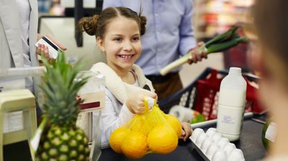 A young girl holds a bag of lemons at the checkout counter of a supermarket.