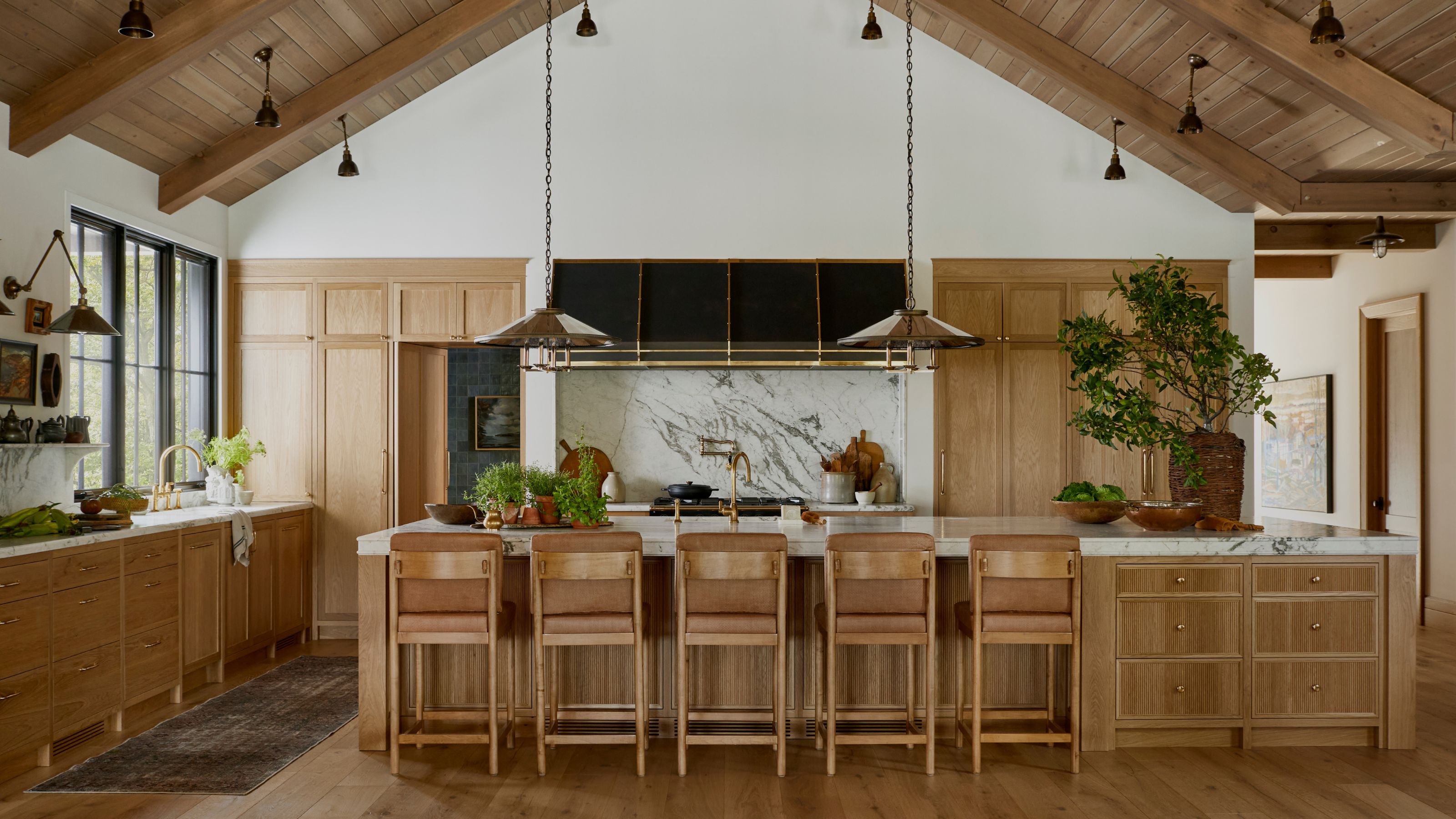 A wide shot of a spacious kitchen with a vaulted wood-beam ceiling, featuring light oak cabinetry, marble countertops, and a large central island with five high-back wooden stools.