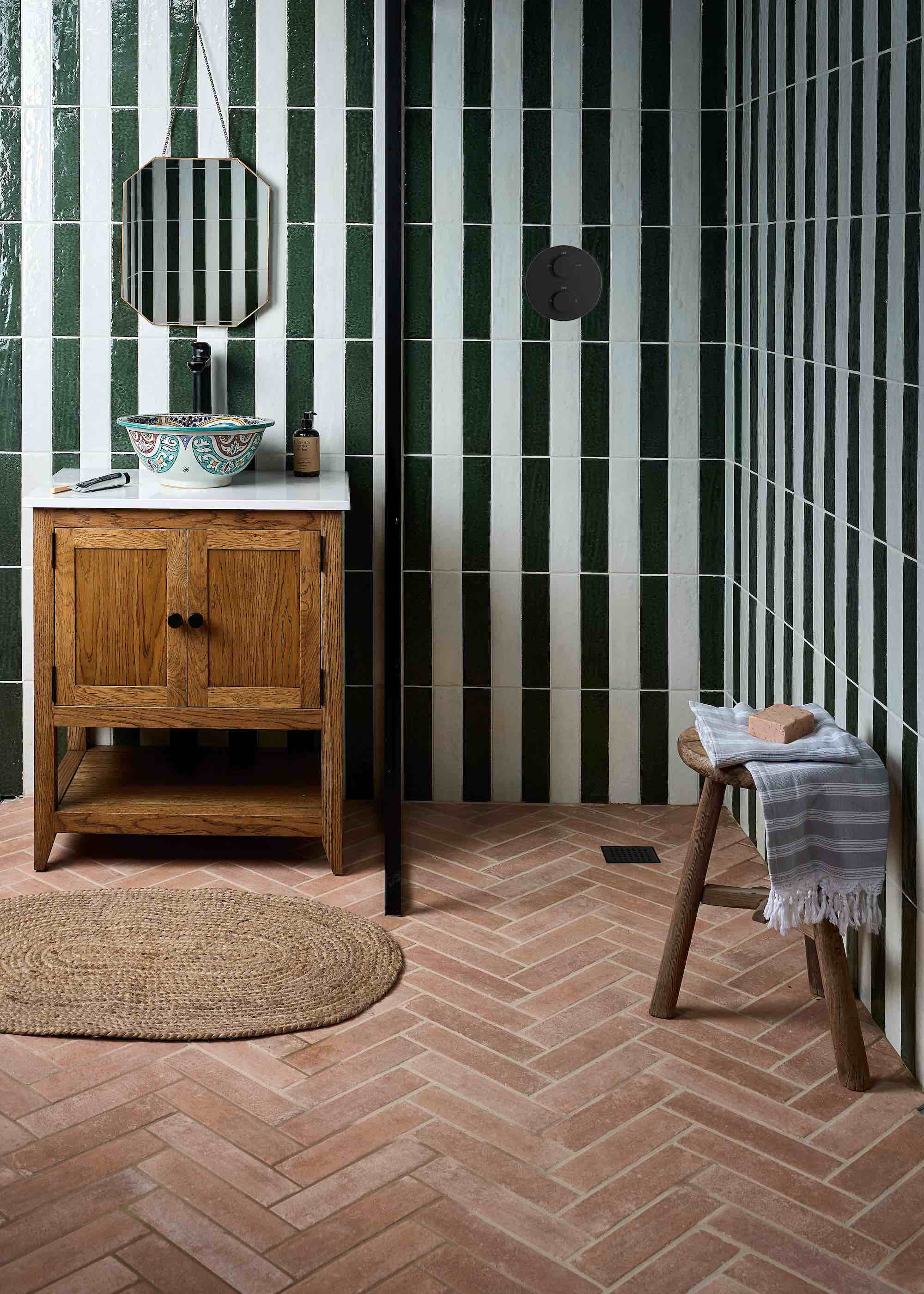 bathroom with herringbone terracotta flooring and striped tiled walls. There is a vanity with a sink on top as well as a hanging mirror.