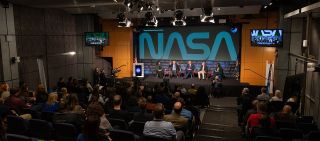 five well-dressed people sit on a stage in front of about 50 seated journalists, with "nasa" written on the wall behind the panelists.