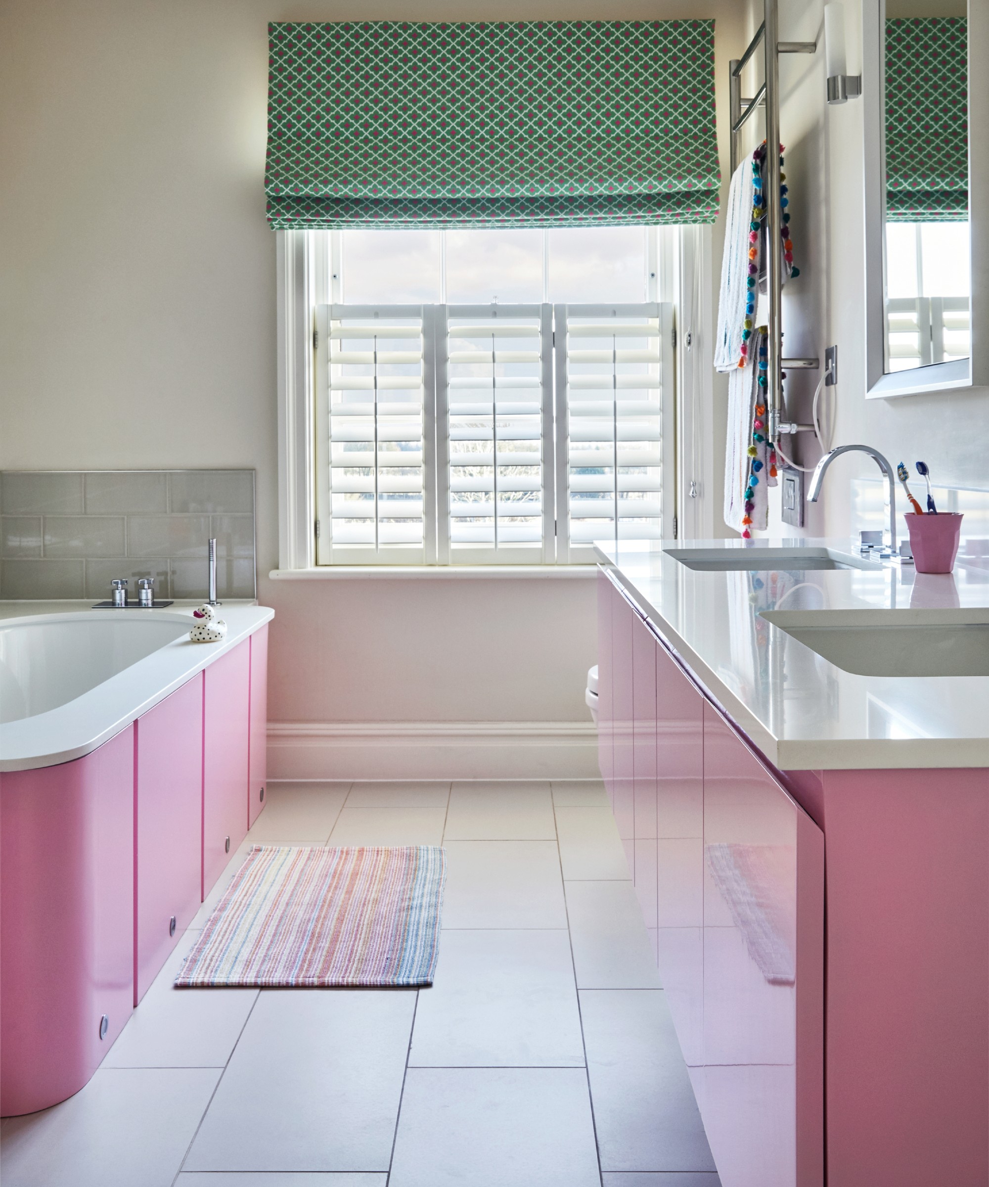 A bathroom with a pink bathtub and matching bathroom cabinets and a patterned green Roman blind on the window