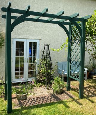 a simple green lattice pergola surrounded by potted and trailing plants, in front of a cream cottage house