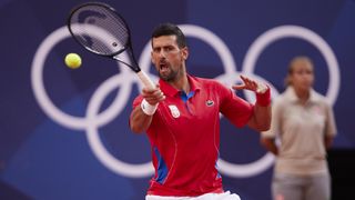 Novak Djokovic, in a red polo shirt and white shorts, plays a baseline shot at the 2024 Paris Olympics.