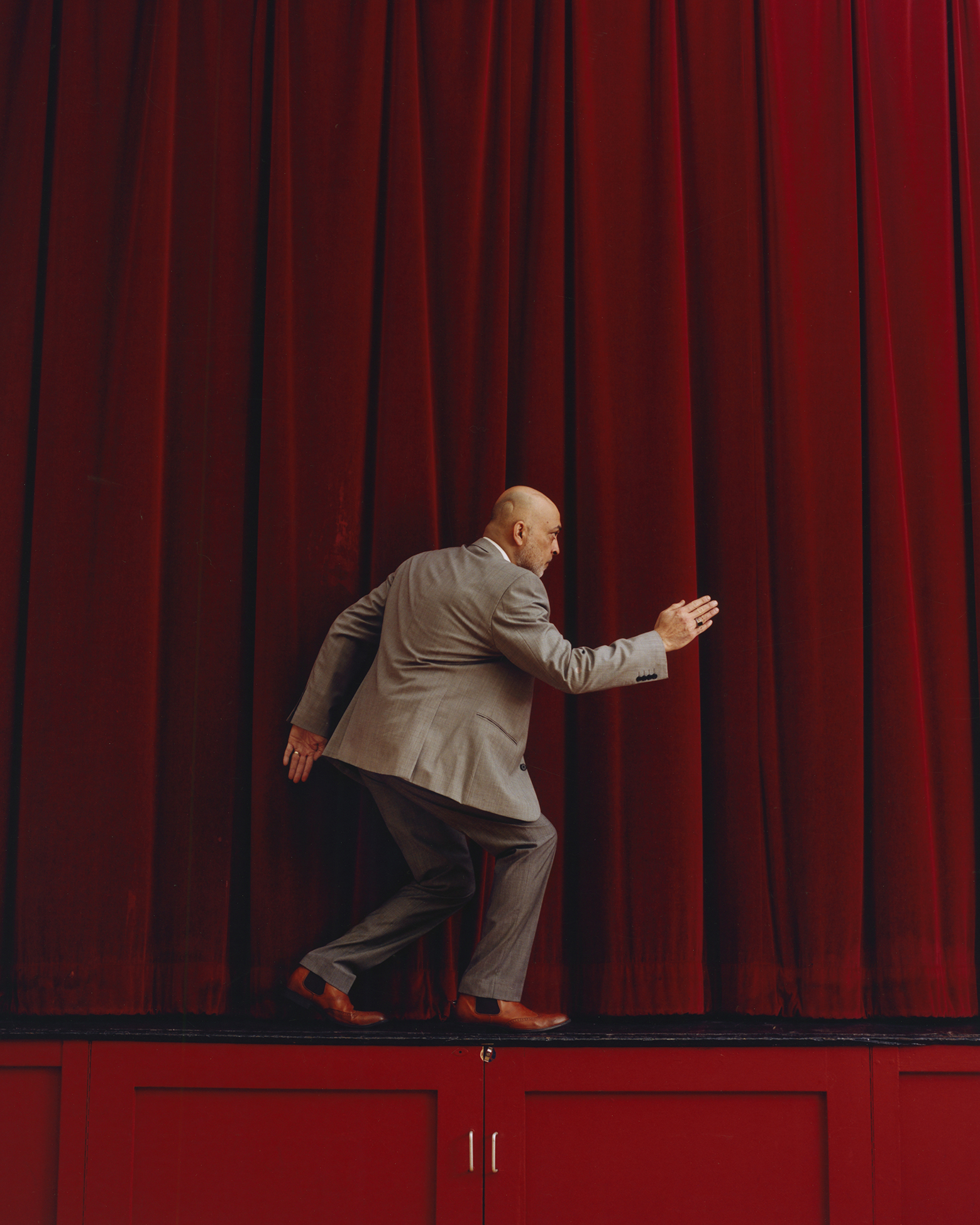 A person in a suit mimics a sneaking pose in front of large red curtains, creating a whimsical and comedic tone. The background is richly colored, enhancing the playful mood