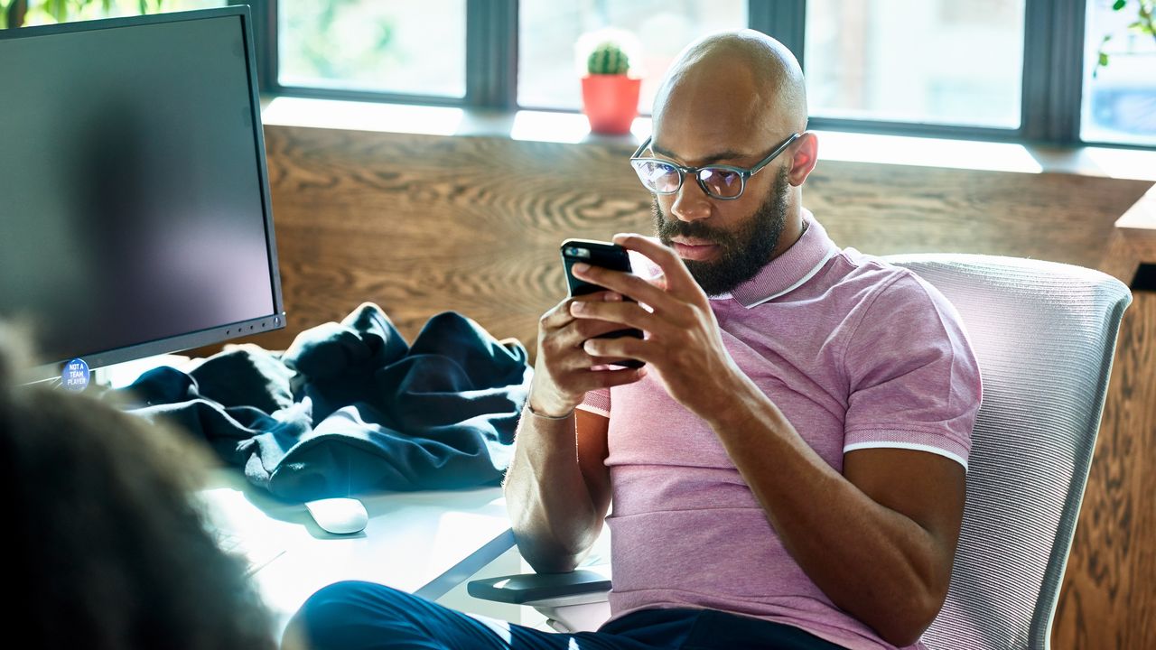 A man sits at his desk in an office and looks at his phone as if he&#039;s researching something.