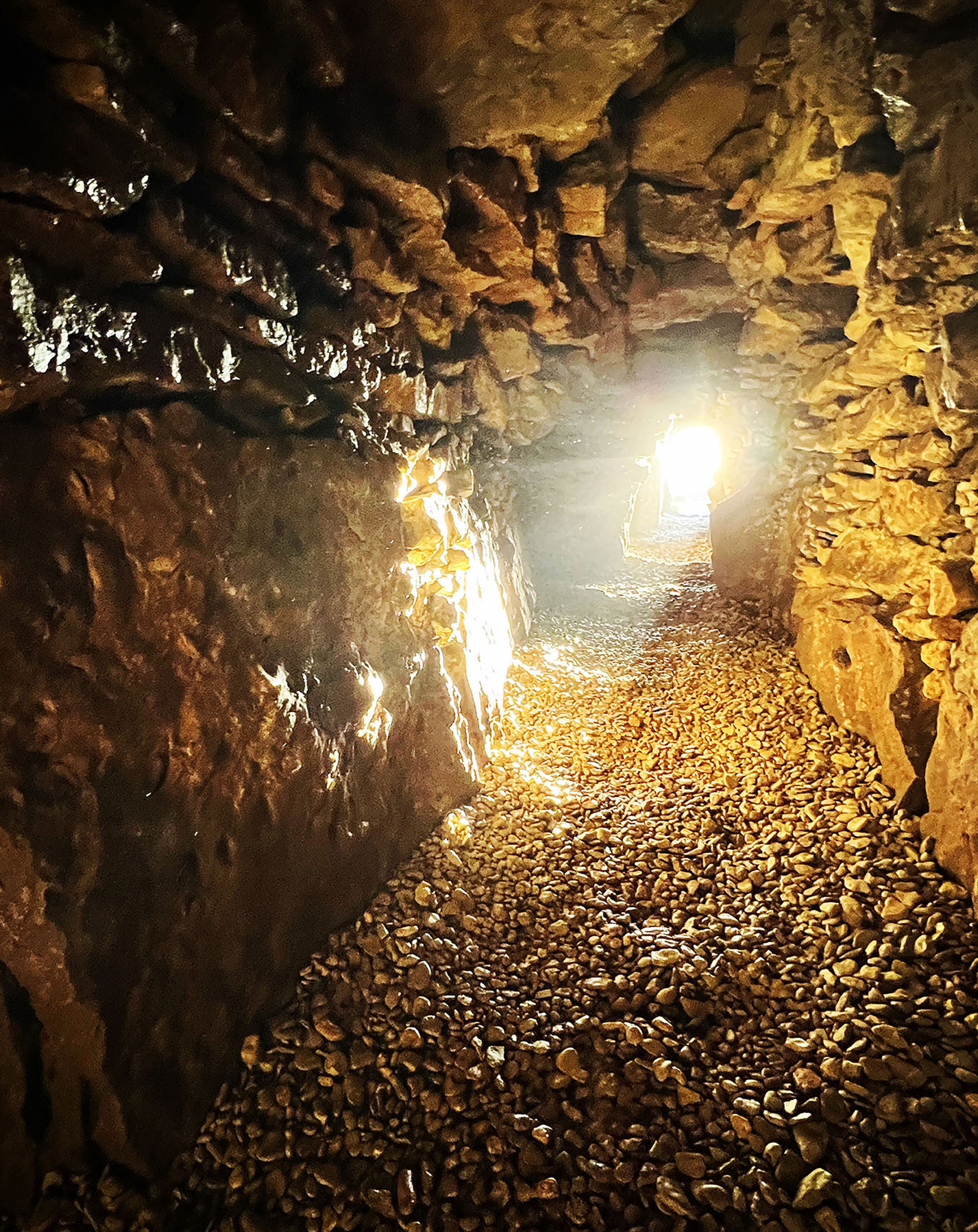a passageway is illuminated with the sun at the far end of the picture. the passageway is ancient, with round stones on the bottom and large stones lining the walls and the ceiling