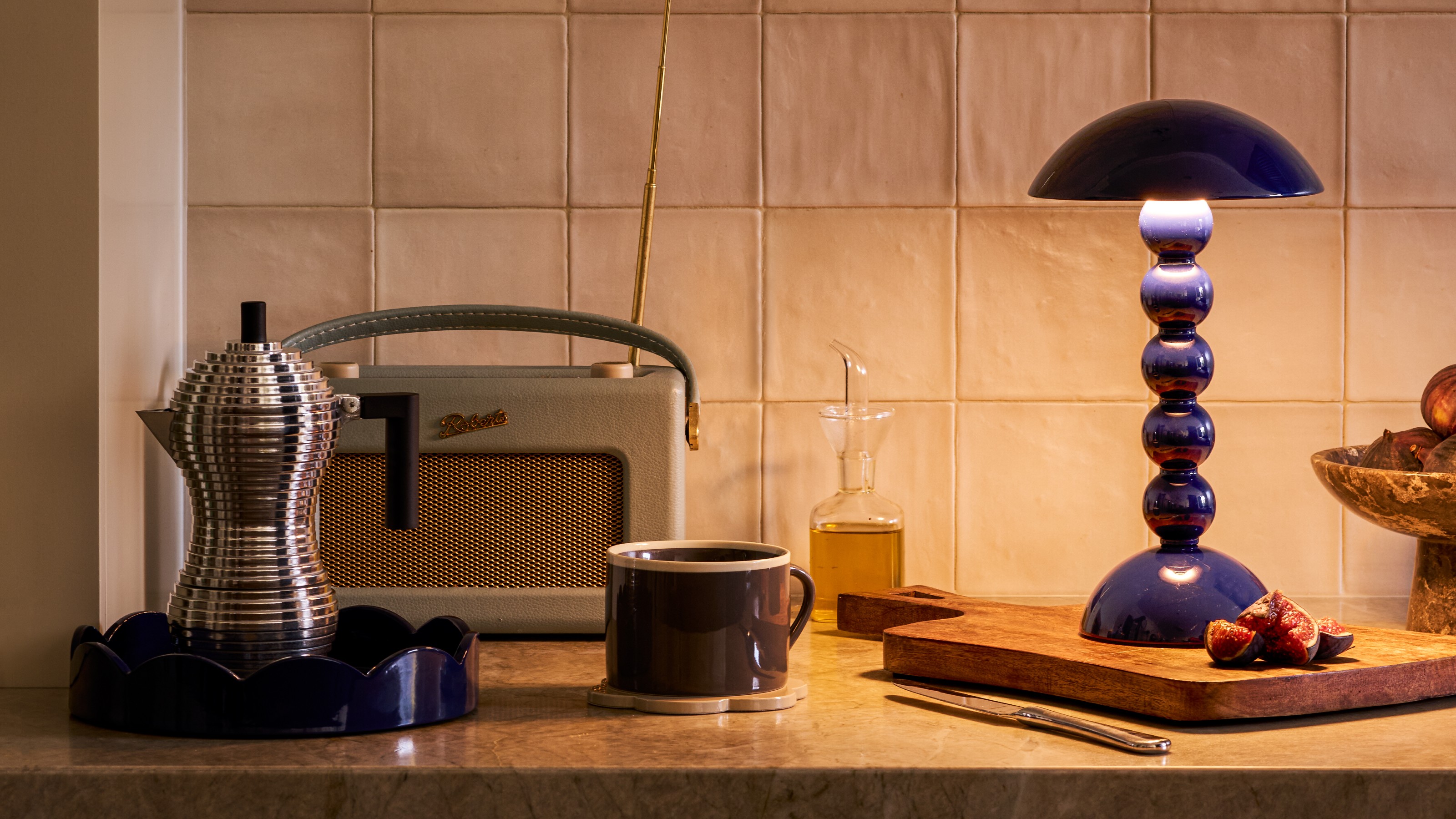 Addison Ross bobbin lamp in navy on a kitchen worktop with a round scalloped matching tray, a Roberts radio and an Alessi espresso maker