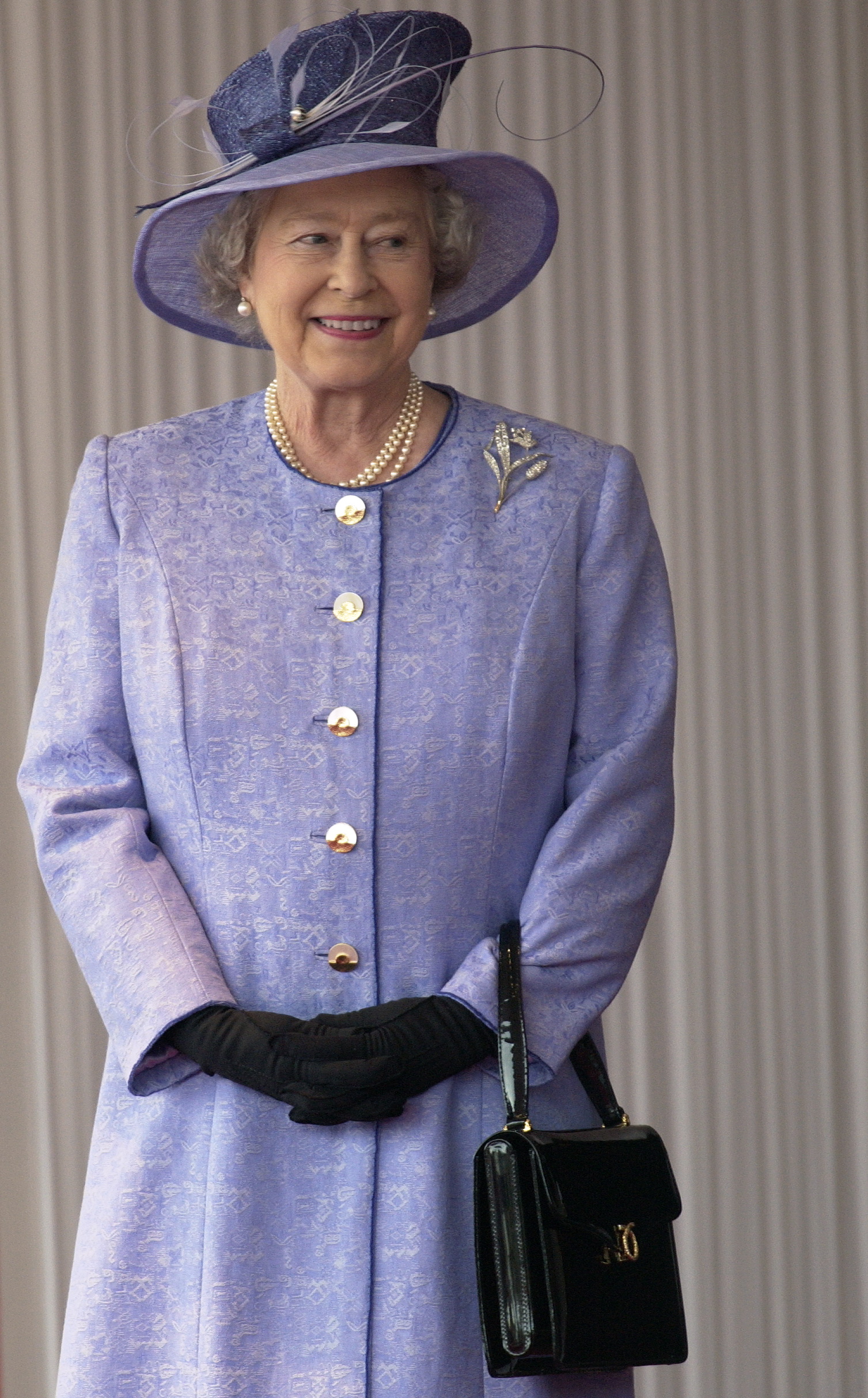 Queen Elizabeth in a lavender coat carries a black Launer bag.