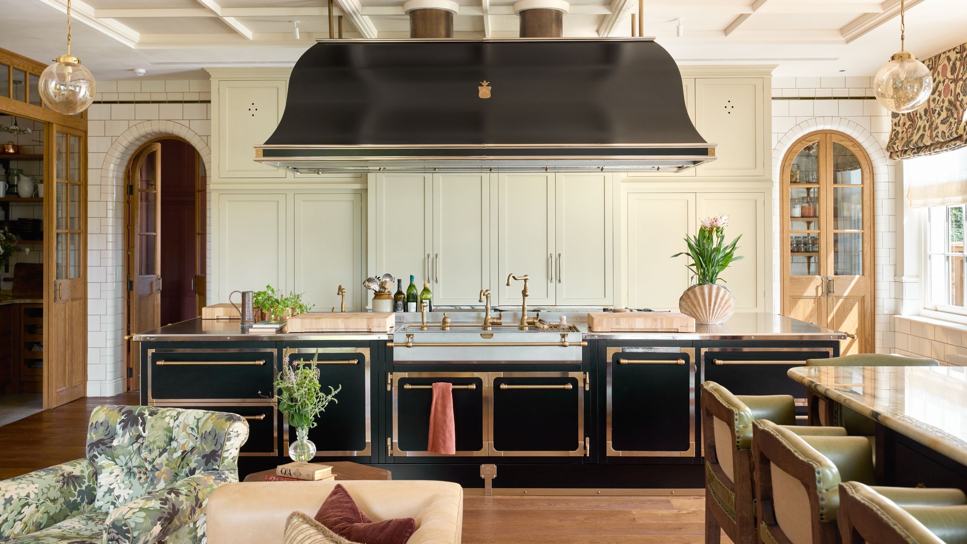 a grand traditional kitchen in Surrey, England with large black and brass island and cook hood, traditional off white cabinetry and arched doors through to pantry spaces