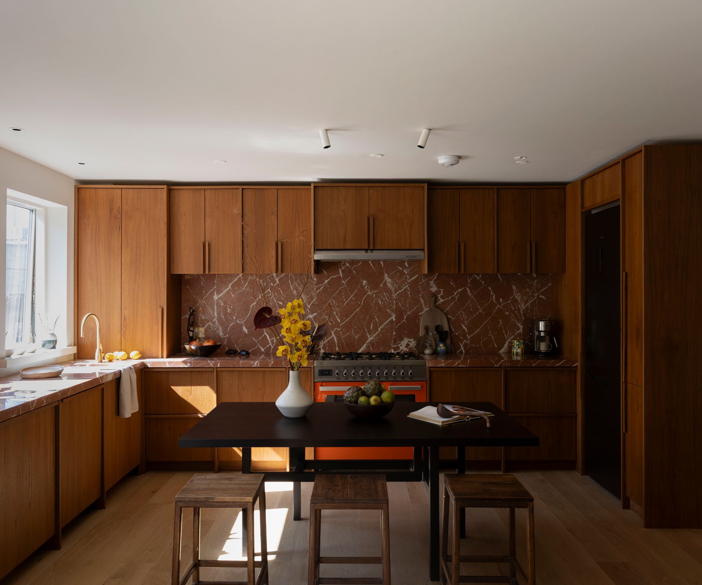 Mid-century modern kitchen with warm wood cabinetry, a dramatic red marble backsplash, and a dark wood dining table with matching stools.