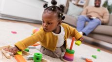 Baby girl playing with toy blocks on the floor at home with her dad seated on a couch in the background