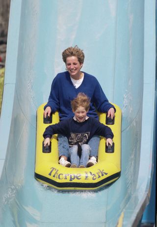 Princess Diana wearing a blue sweater and jeans sitting in a yellow raft with Prince Harry on a water slide