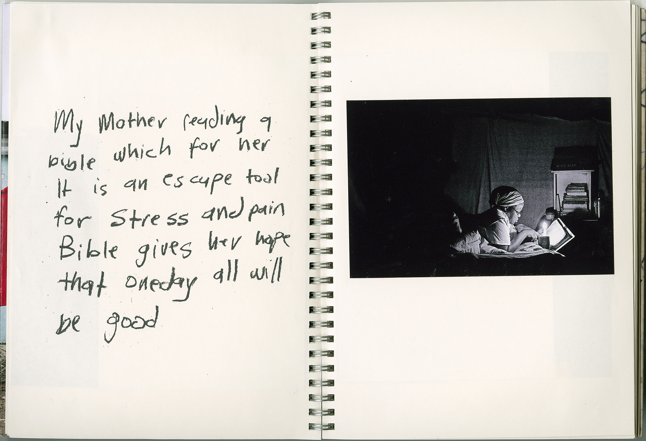 Color photograph of a notebook with an image of a woman reading a book by candlelight on the right side. On the left, the words "My mother reading a bible which for her is an escape tool from stress and pain. The Bible gives her a sense of hope that one day all will be good." are written