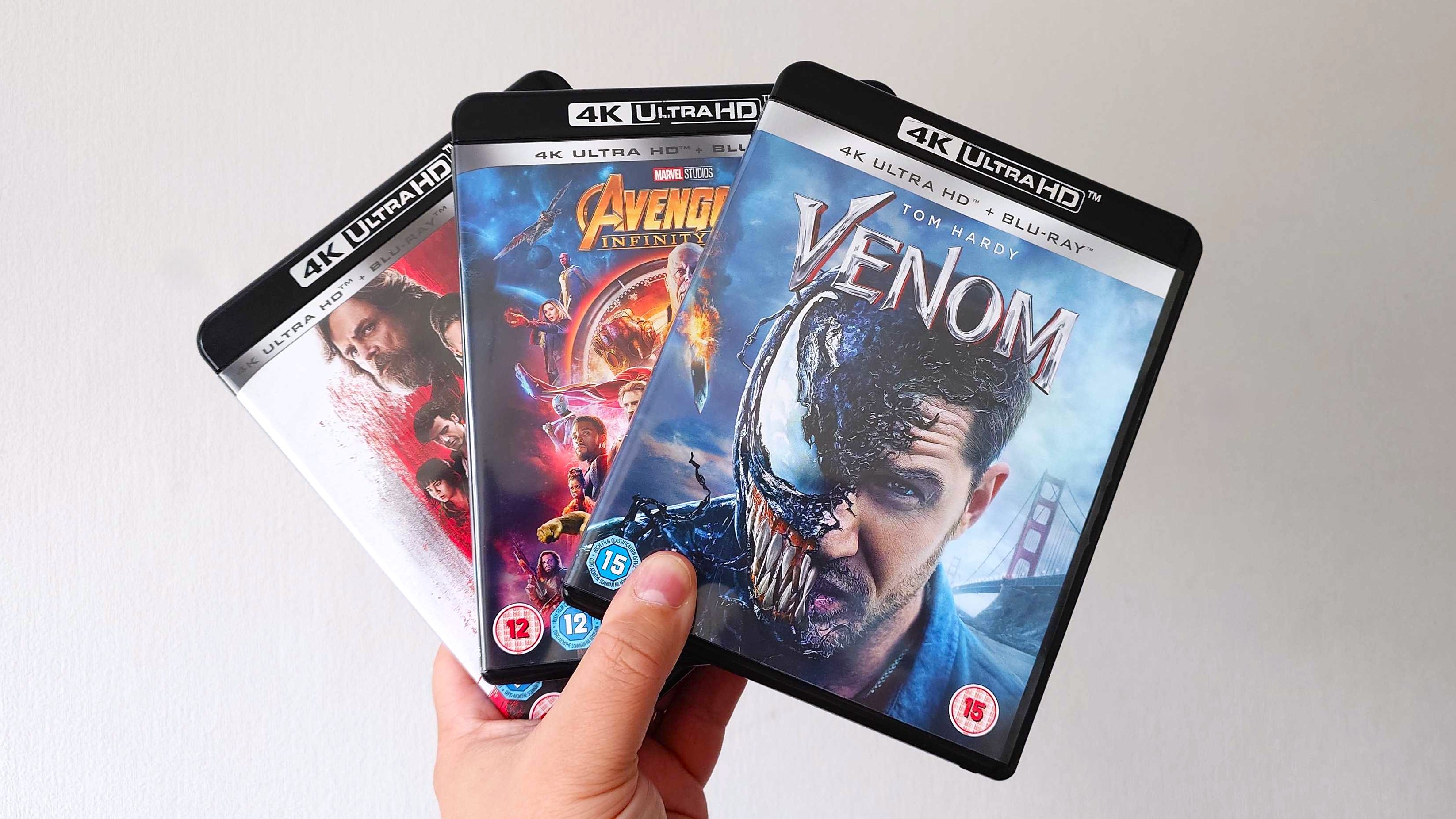 A hand holding Venom, Avengers Infinity War, and Star Wars The Last Jedi 4K Ultra HD blu-ray discs in their cases.