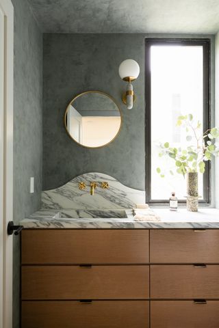 A bathroom with green lime wash walls, a circular mirror, and a marble and wood vanity