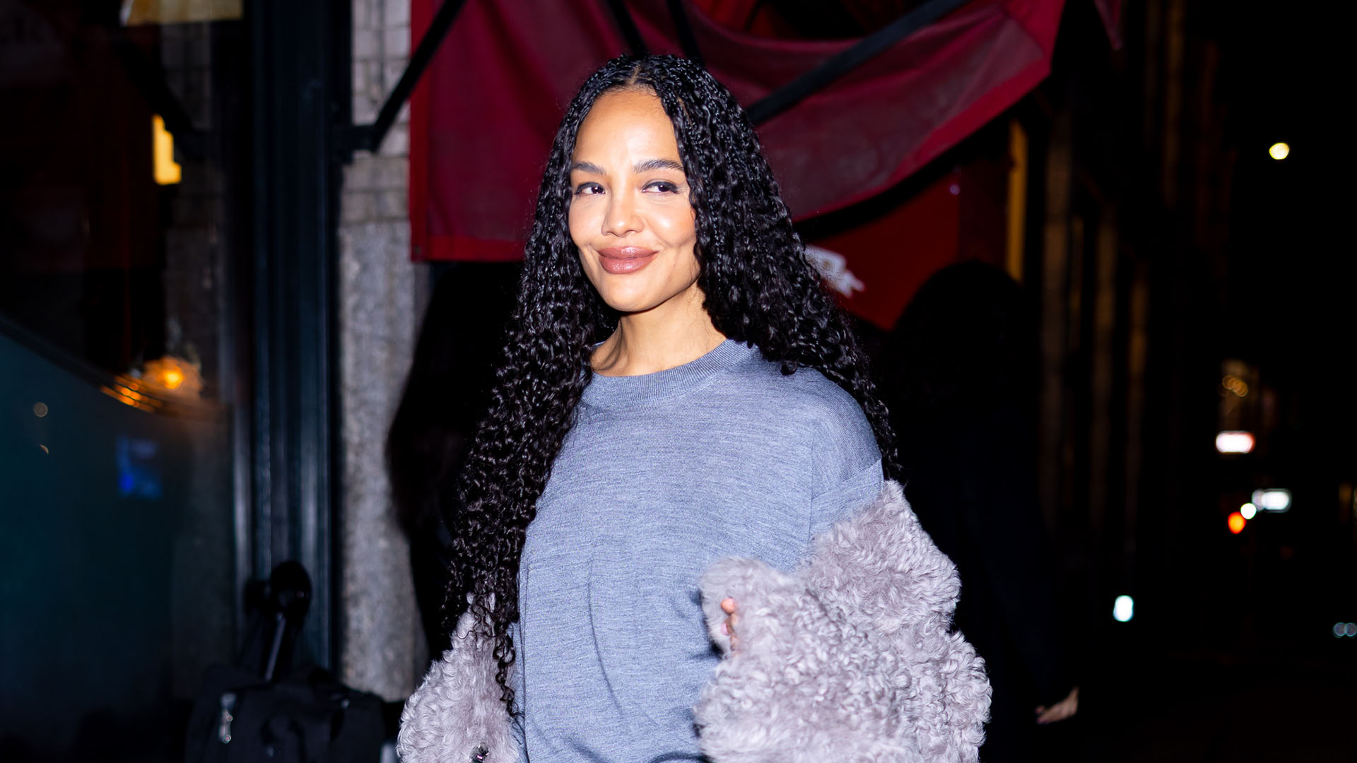 Tessa Thompson wearing a gray sweater, gray fur jacket, black satin skirt, and black boots