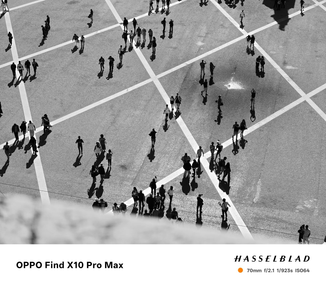 Black-and-white overhead view of people and long shadows crossing a geometric city square.