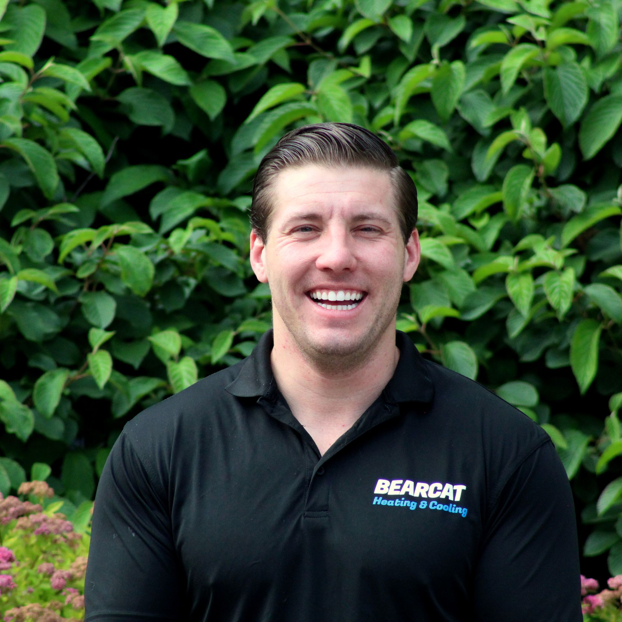 A headshot of Alex Marsh wearing a black top in front of a green and leafy bush