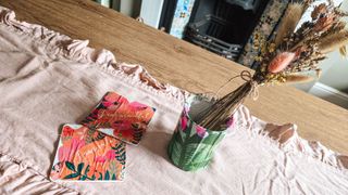 A wooden dining table with a light pink frilled table runner with two coral decoupage mats and a green decoupage tin with dried flowers in it