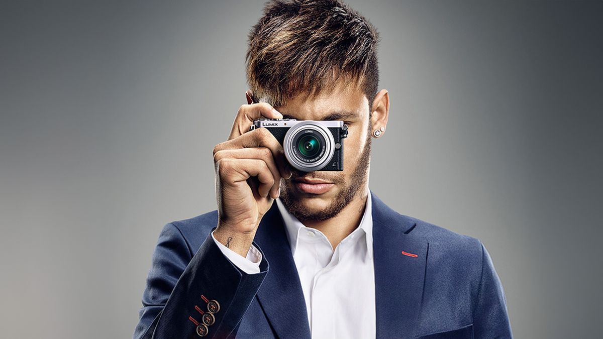 A man in a blue suit, with a white shirt, holding a Panasonic Lumix GM1 camera up to his eye, obscuring his face