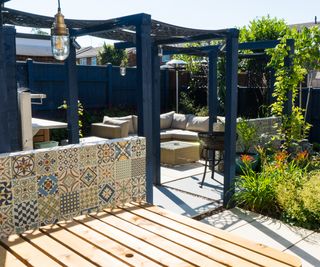 Pergola covered seating area with light coloured paving and decking as well as a comfortable looking corner sofa