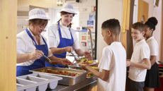 Three school children queuing for school lunch, which is being served by two dinner ladies