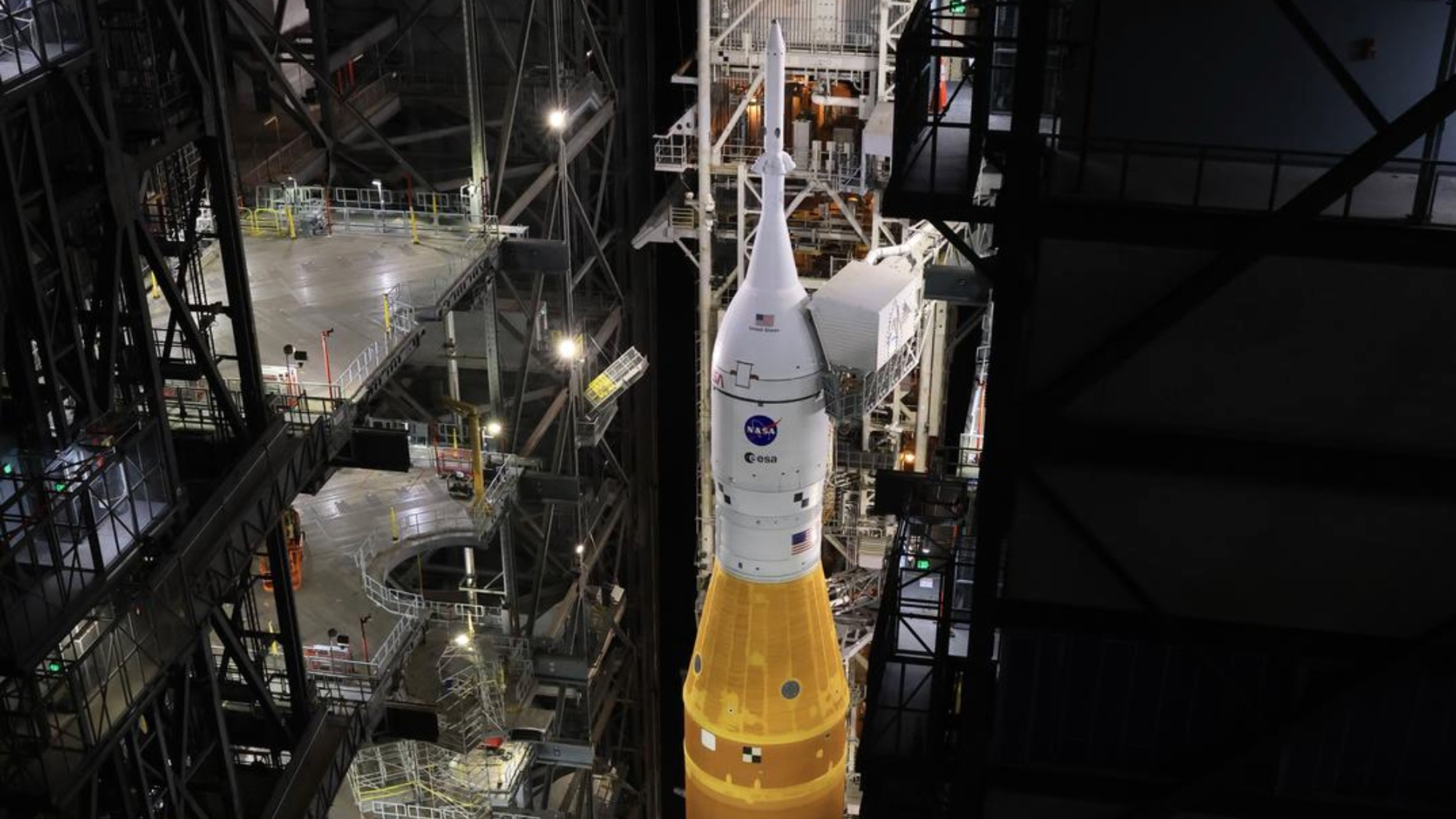 a white space capsule sits atop an orange rocket inside a cavernous hangar at night.