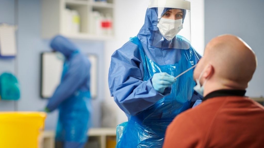 health care worker in protective clothing, mask and face shield administering a nasal swab test to a male patient in the foreground