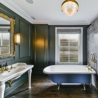 a bthroom with dark green walls and dark wooden floorboards, a blue and white freestanding rolltop bathtub, a large white vanity sink with a gold framed mirror above it