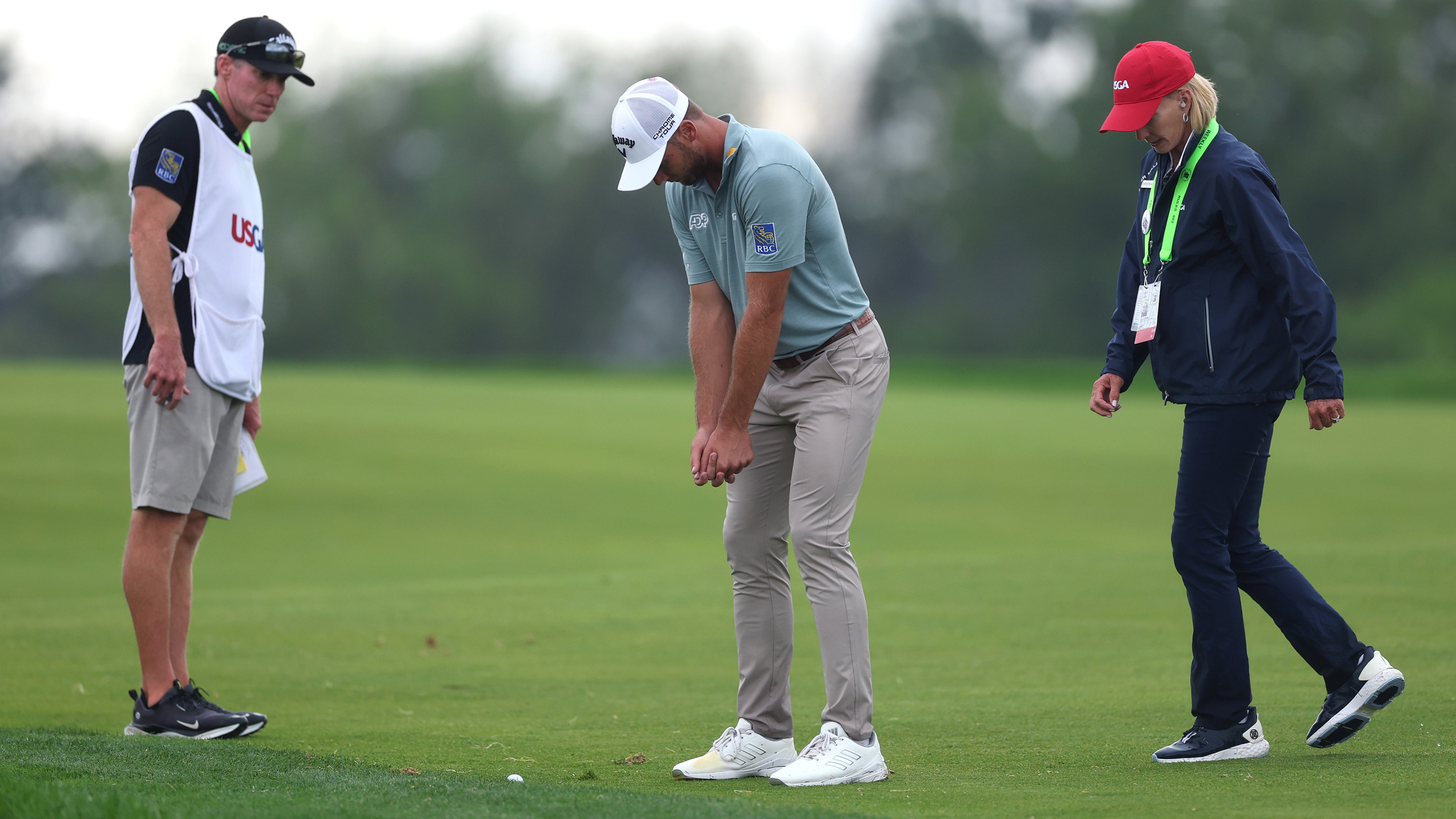 Sam Burns poses in his swing stance while discussing with a US Open official as to whether he should receive a free drop from casual water at Oakmont