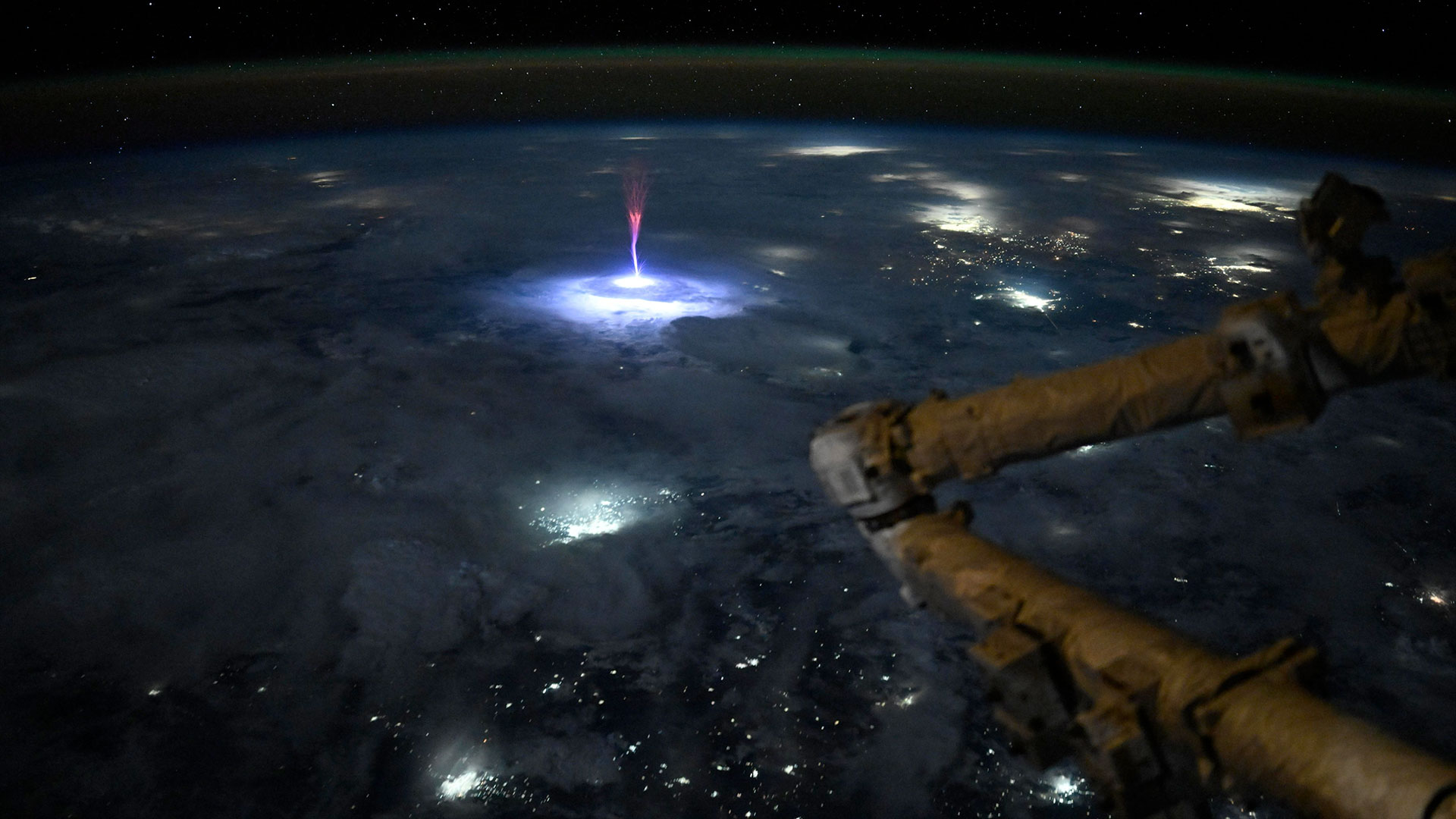 a bright red spark jets up from above the clouds looking down at lightning storms at night from a space station