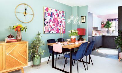 Duck egg blue open plan kitchen and dining room with orb pendant light, abstract artwork on wall, industrial dining table and blue velvet chairs