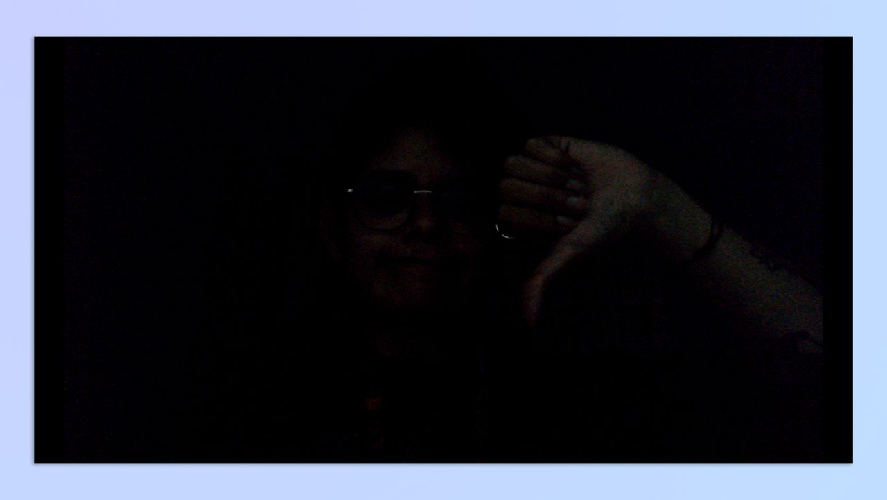 A woman with short hair sitting in a dark room, holding up her hand in the shape of a thumbs-down