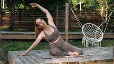 A woman practices Pilates outdoors on an exercise mat. Her legs are bent to the side and she is leaning over, one arm bent over her head.