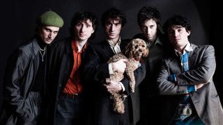 A portrait of Fat White Family stood in front of a dark background. One member is holding a puppy