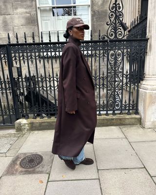 @nlmarilyn wears a long brown coat, light blue jeans, brown suede loafers and a light brown baseball cap